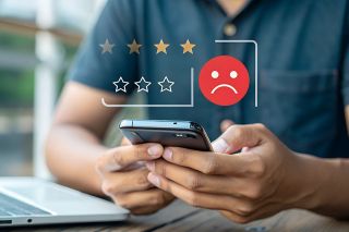 Foto: Mann bedient Smartphone, darüber negatives Smiley und schlechte Sterne Bewertung